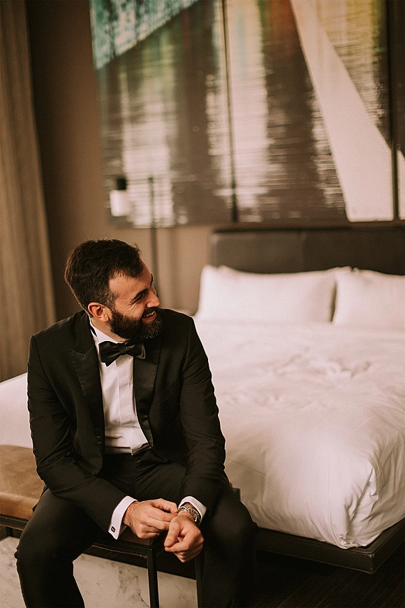 Groom portrait of a bearded groom getting ready in a black tuxedo and bow tie, sitting on a bench in a hotel bedroom by the bed