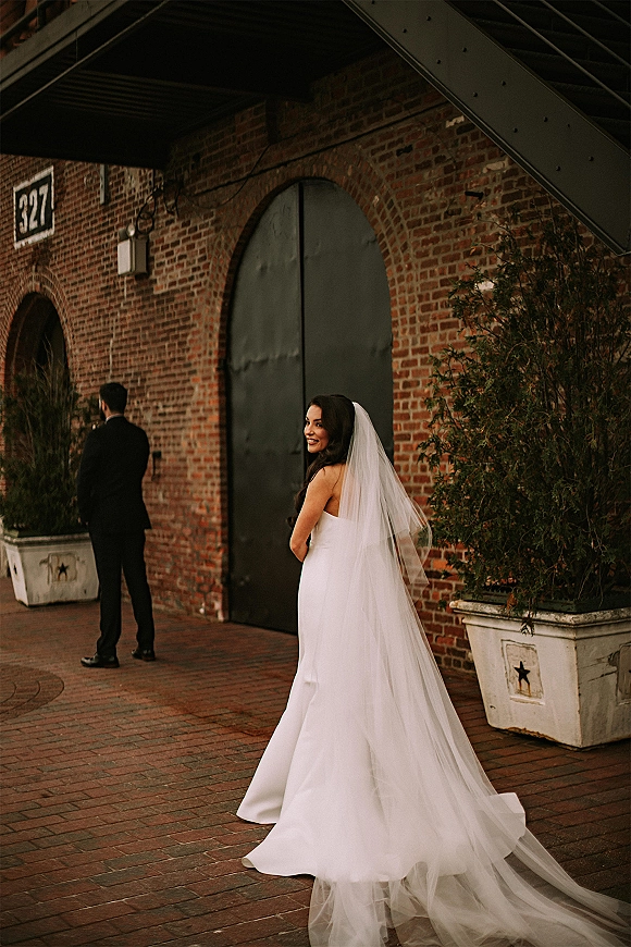 First look moment as bride approaches groom from behind in a long veil, brick courtyard setting with arched metal door and stairs