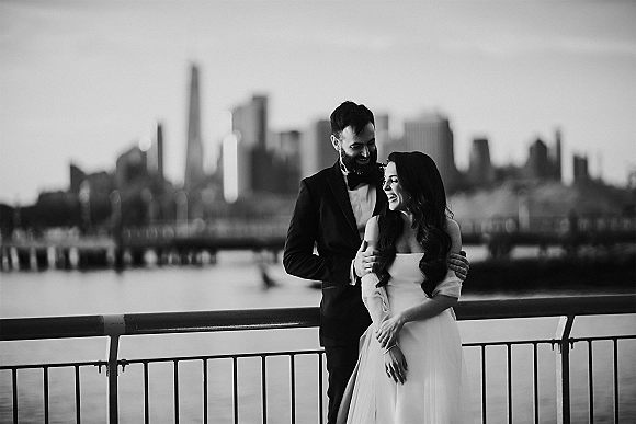 Couple portrait, black and white wedding portrait of bride and groom laughing as he hugs her by a waterfront railing with city skyline and bridge behind them