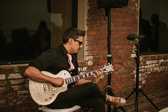 Wedding musician in sunglasses playing electric guitar with a pink tie by a microphone stand, set against a brick wall in an urban venue