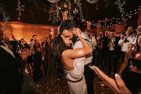 Wedding kiss moment as newlyweds dip-kiss on the reception dance floor, surrounded by cheering guests with drinks under string lights