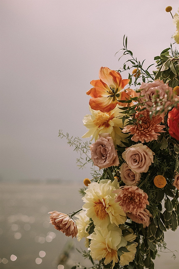 Wedding bouquet with orange and blush bouquet blooms of tulips, roses, and dahlias on a flower stand by a sunlit ocean horizon