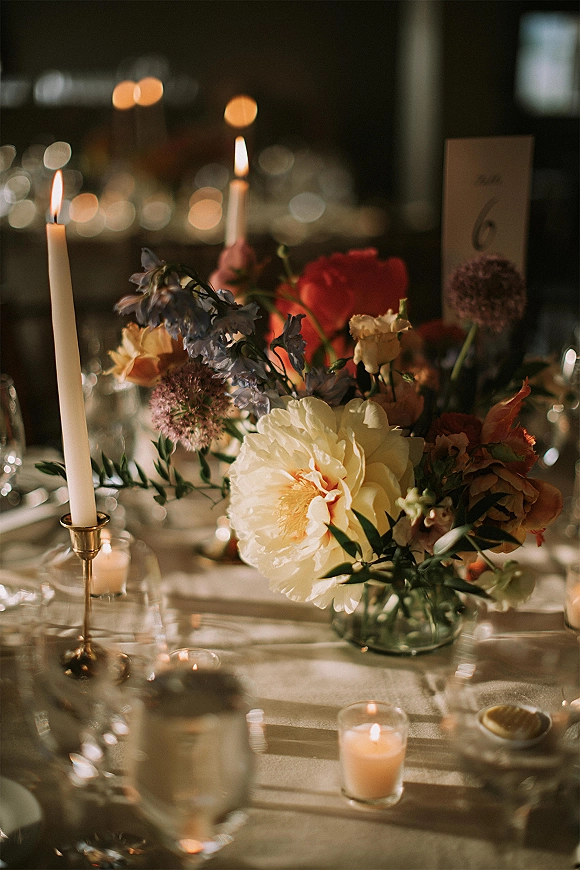Reception tablescape with wedding table centerpiece, taper candles in brass holders, floral arrangement and table number in a dim room with bokeh lights