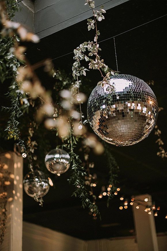 Disco ball decor with wedding disco balls hanging among greenery garlands and string lights from dark ceiling beams above reception space