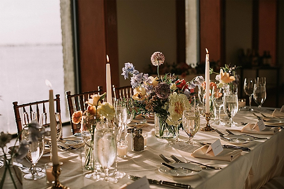 Reception tablescape with wedding table centerpiece of brass candlesticks, white taper candles, florals, and place cards in window light