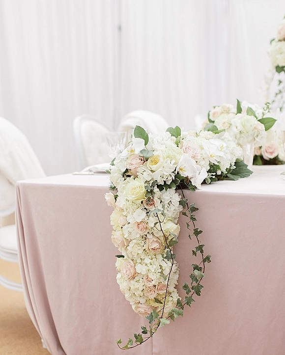 Reception head table with cascading table floral of roses, orchids, and white hydrangea on blush linen, set against white drapery
