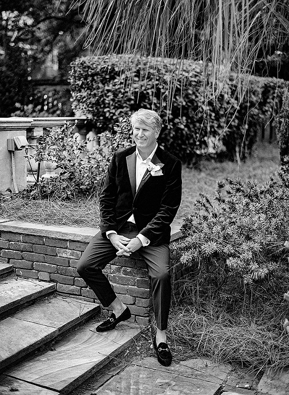 Groom portrait of a man seated on brick steps in a velvet tuxedo jacket with a white boutonniere, wristwatch, and loafers in a garden setting