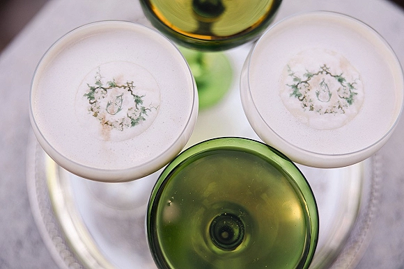 Signature cocktails in coupe glasses with foam topping and botanical garnish, arranged on a silver tray over a white surface
