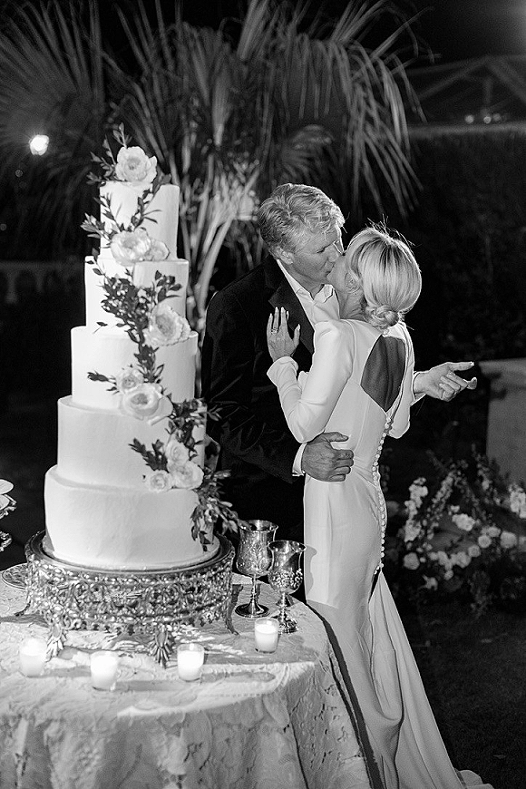Wedding kiss beside a three tier wedding cake, bride in long sleeve gown and groom in dark suit at candlelit garden reception at night
