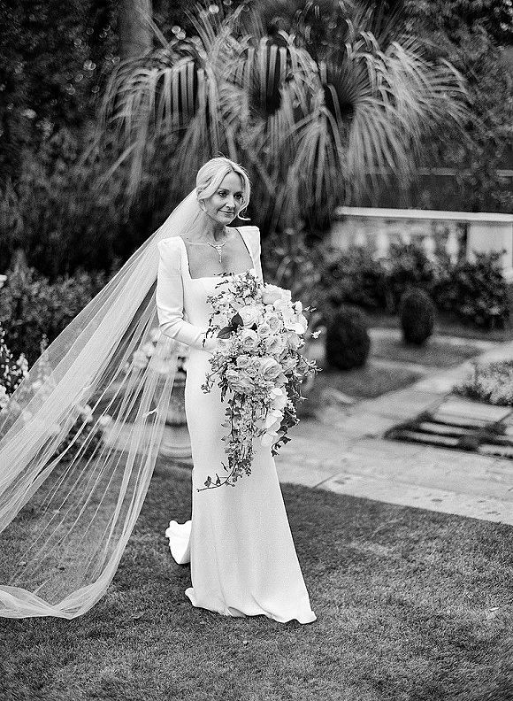 Bridal portrait of a bride holding bouquet with a long cathedral veil flowing over a garden lawn beside palm fronds and stone steps