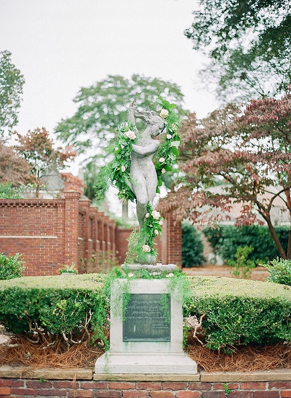 Wedding floral wreath with roses and greenery garland draped on a stone statue pedestal along a hedge-lined garden walkway near brick wall