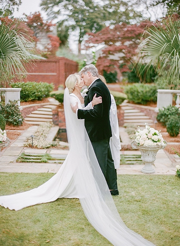 Wedding kiss portrait of bride and groom kissing, her long veil flowing as they embrace on stone steps in a garden by a brick wall