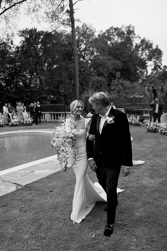 Wedding couple portrait of bride and groom walking, she holds a cascading bouquet, with veil and tuxedo by a poolside garden lawn
