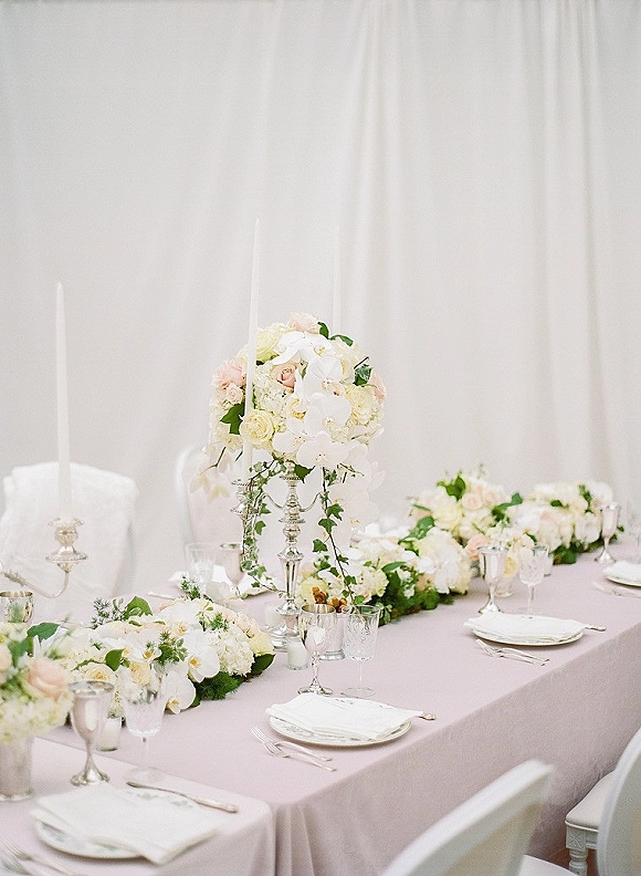 Reception tablescape with soft pink tablecloth, white floral garland, tall centerpiece, silver candlesticks and taper candles before draped backdrop