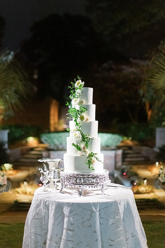Wedding cake with white buttercream in a five tier wedding cake design, wrapped in greenery garland on an ornate silver stand outdoors