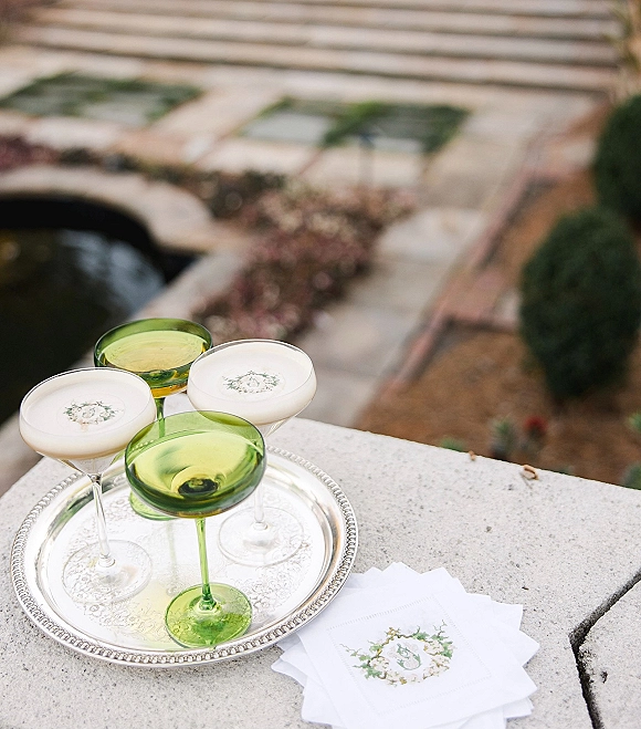 Wedding cocktails on a silver tray with green coupe glasses and foam topped drinks, paired with monogram napkins by garden steps