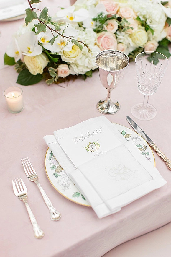 Reception tablescape with blush wedding table setting, rose-and-orchid centerpiece, single votive candle, and silver goblet beside crystal glassware