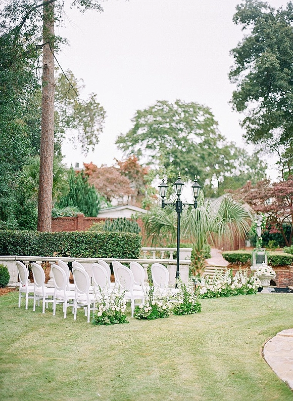 Ceremony setup with white ceremony chairs lining a garden aisle, framed by greenery garland and ground florals by a brick wall backdrop