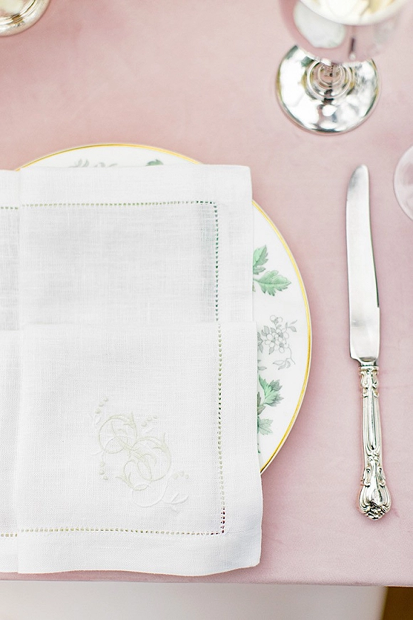 Reception place setting with monogrammed white linen napkin on floral china and gold-rim plate, beside a wine glass on blush pink tablecloth