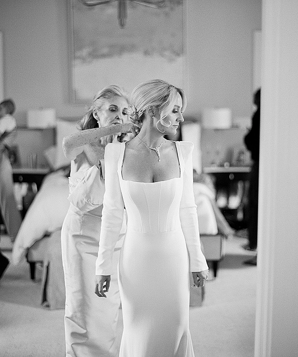 Bridal getting ready as mother helps button wedding dress with long sleeves and square neckline in a hotel suite, drop necklace glinting