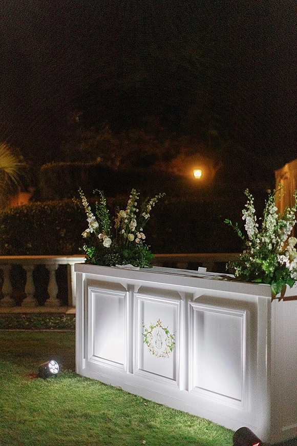 Wedding bar setup with a white paneled front and monogram emblem, topped with floral arrangements and greenery on a lawn at night