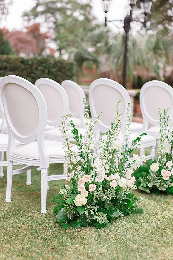 Ceremony aisle decor with aisle floral arrangements of garden roses, greenery and blush blooms beside white chairs on a grass lawn