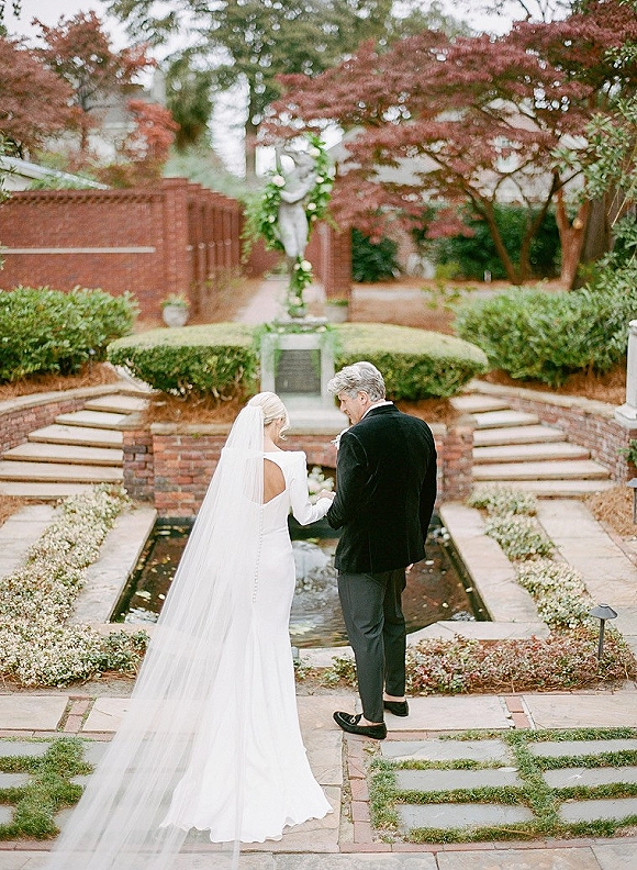 Couple portrait of bride and groom back view holding hands, her long veil trailing by a floral arch on garden stone steps near a reflecting pool