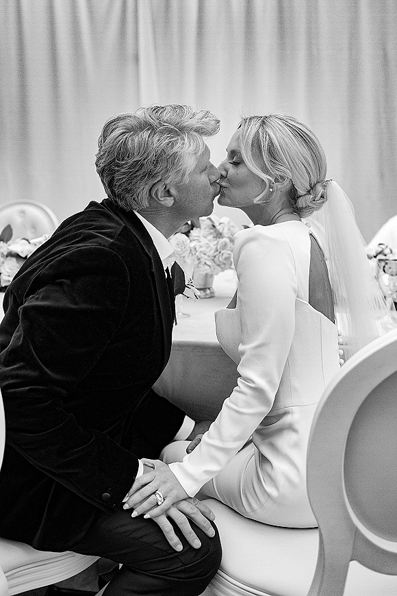 Wedding kiss portrait in black and white wedding photo as seated newlyweds kiss at a reception table with draped fabric backdrop and florals