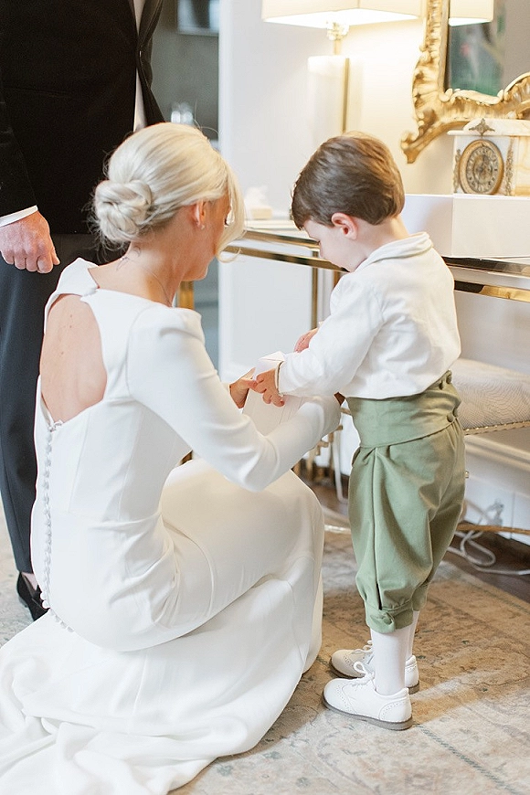 Getting ready moment as a bride with ring bearer kneels to help him dress, gown buttons showing beside groom in black suit indoors