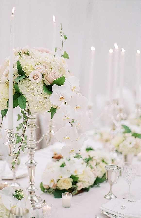 Reception tablescape with a wedding table centerpiece of roses, hydrangea and orchids around silver candelabras and white taper candles on linen