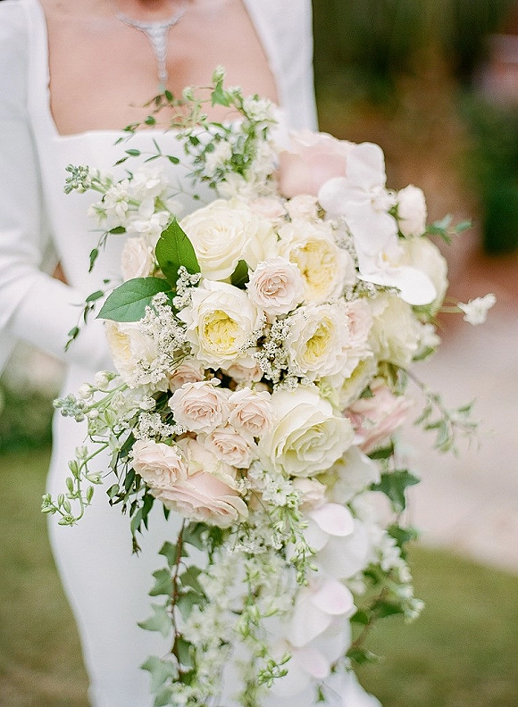Bridal bouquet with a white rose bouquet of blush roses, orchids, and greenery held against a bridal dress in an outdoor garden walkway