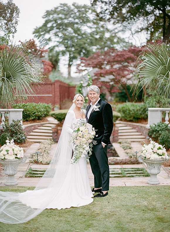 Couple portrait of bride in strapless gown with long veil and cascading white rose bouquet beside groom in tux on stone steps in garden
