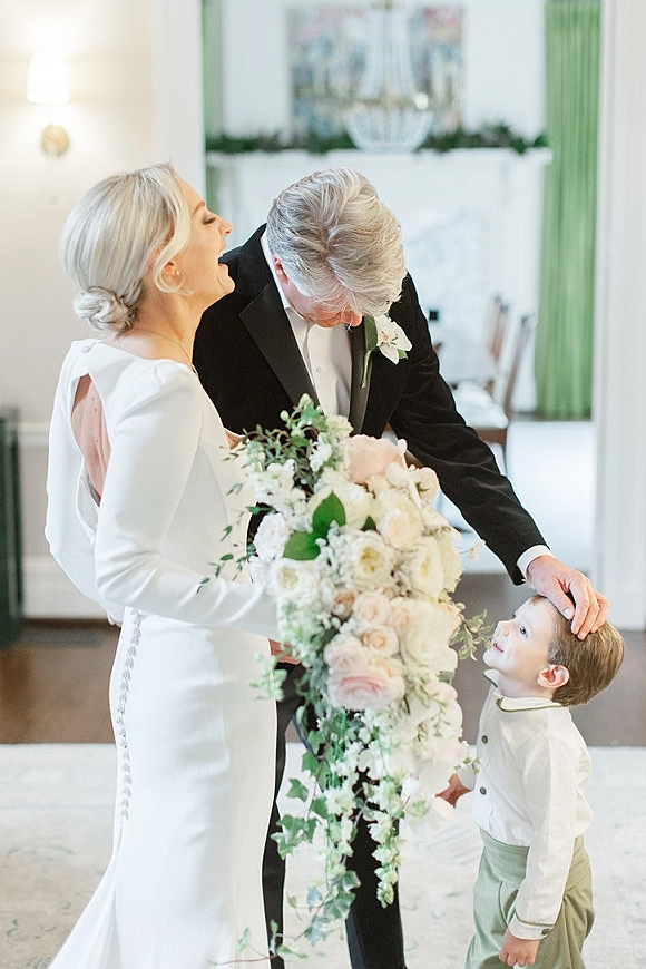 Wedding family portrait of bride groom and child indoors, bride laughing in long sleeve dress holding cascading bouquet beside tuxedo groom
