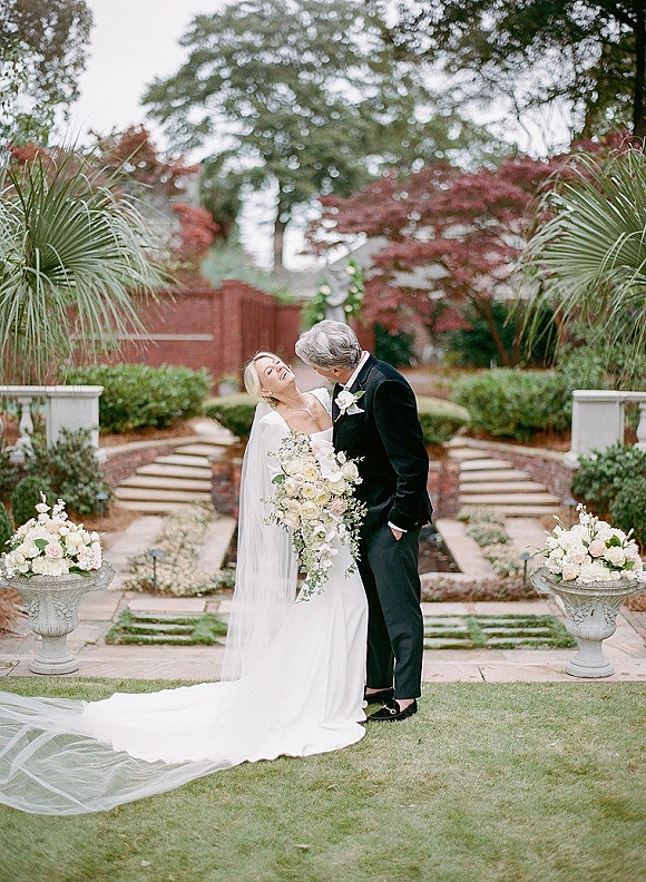 Couple portrait of bride and groom laughing, bride holding bouquet with long veil and train on garden steps by brick wall