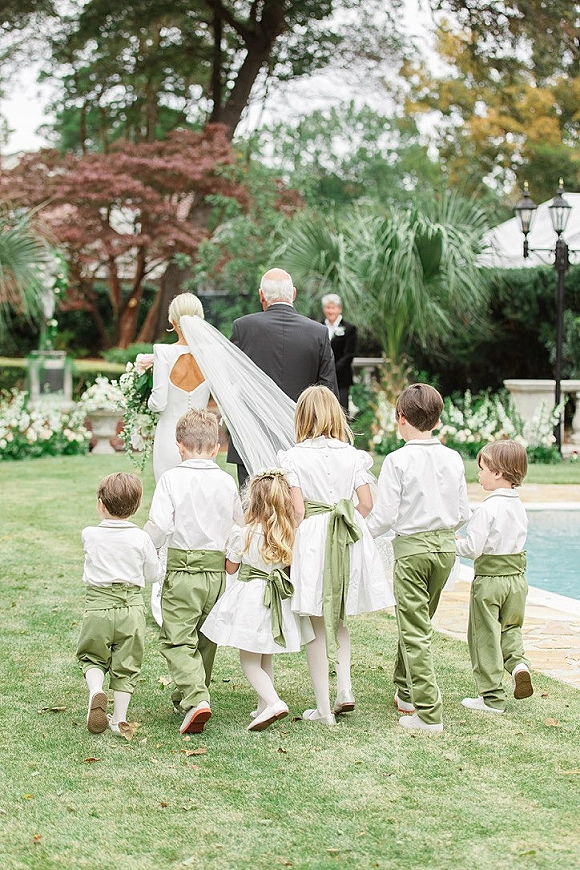 Wedding processional with bride walking down aisle in a long veil, holding bouquet, children following on a garden lawn by the pool.