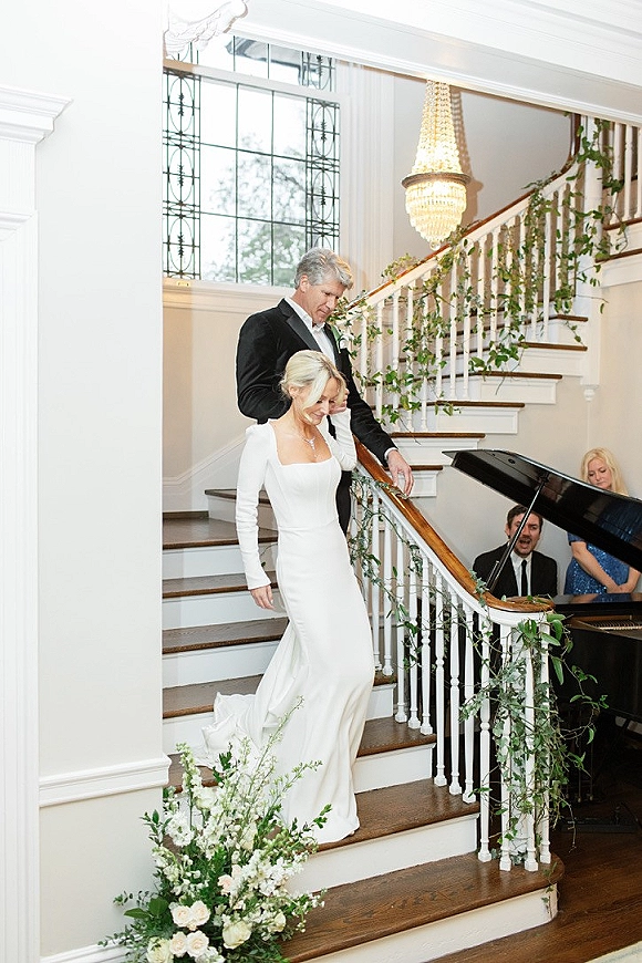 Bride and groom entrance down a wedding staircase entrance, groom guiding her past greenery garland and chandelier in an elegant foyer