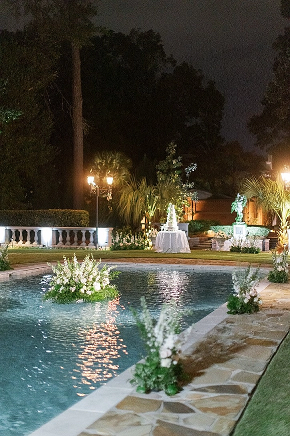 Poolside wedding decor with floating floral centerpieces and white rose arrangements beside a cake table, softly lit in a garden at night