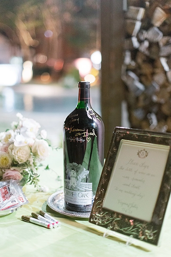 Wedding guest book wine bottle guest book on a silver tray with framed sign, markers, and blush florals, set before bokeh lights and stacked firewood