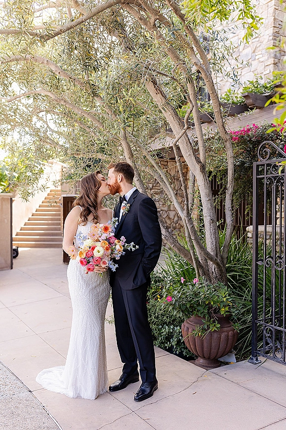 Wedding kiss portrait of bride and groom kissing, bride holding a colorful bouquet in a lace dress by a wrought iron gate and stone steps