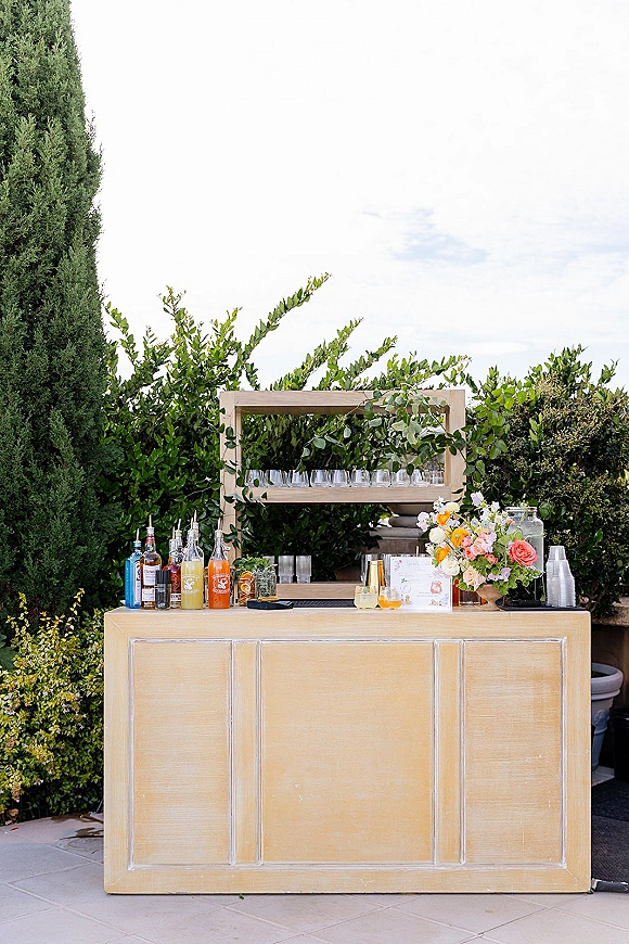 Wedding bar setup on a wood bar with outdoor wedding bar shelf of liquor bottles, glassware, and greenery garland on a patio