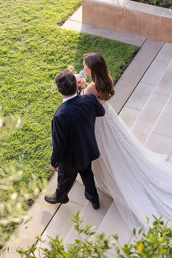 Couple portrait of bride and groom walking down stone steps, her long wedding dress train trailing behind as she holds a bouquet amid greenery