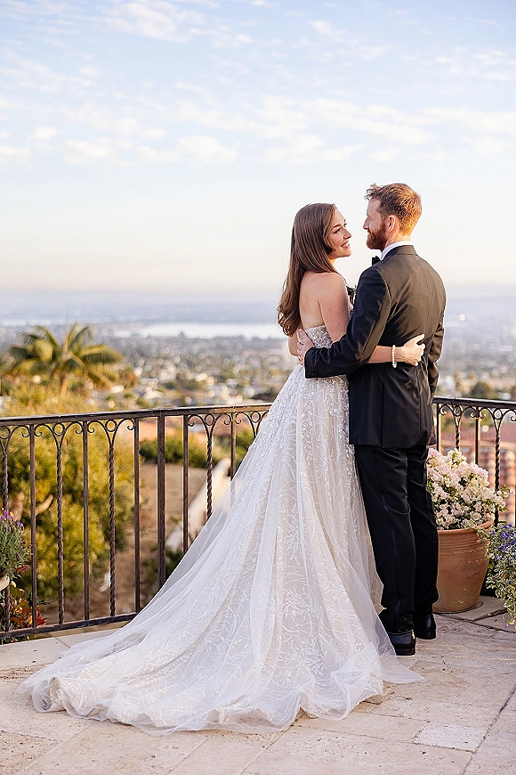 Couple portrait of bride and groom embrace on a terrace patio, her veil and lace dress train flowing by wrought iron railing, ocean and city skyline behind