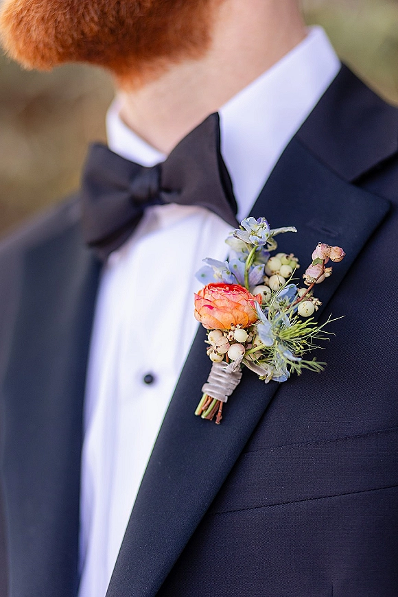 Wedding boutonniere with orange ranunculus and blue blooms pinned to a black tux lapel, ribbon-wrapped, against blurred greenery backdrop