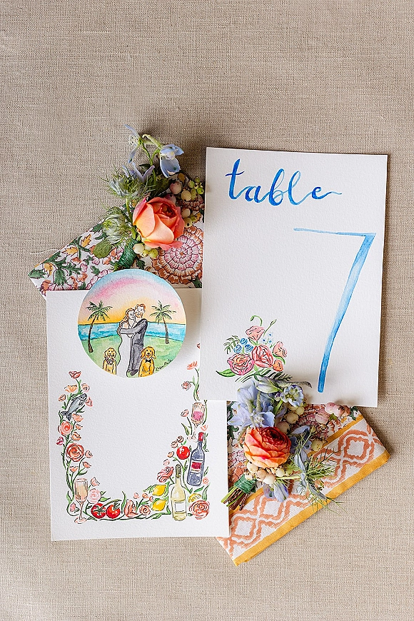 Wedding stationery suite with illustrated card and calligraphy table number, accented by orange ranunculus on a linen fabric backdrop