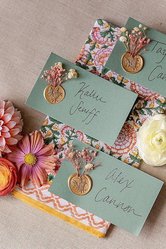 Wedding place cards with calligraphy place cards on green cardstock, sealed with gold wax and dried flowers, styled on a linen tablecloth