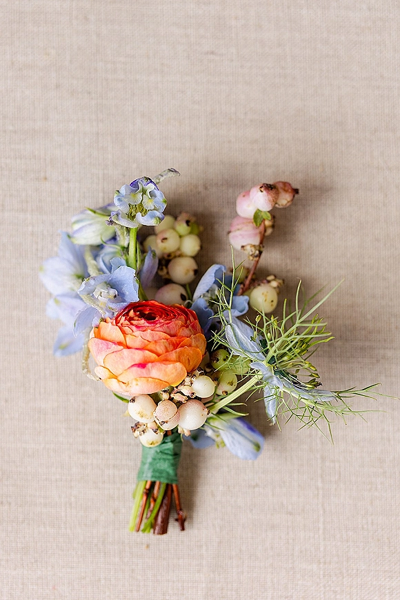 Boutonniere featuring orange ranunculus and blue delphinium with white berries and greenery, wrapped stems on a linen backdrop flat lay