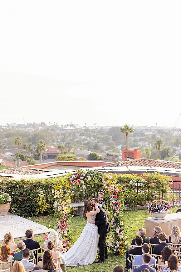 Ceremony kiss as bride and groom embrace under a floral arch, guests seated on a lawn terrace with palm trees and cityscape beyond