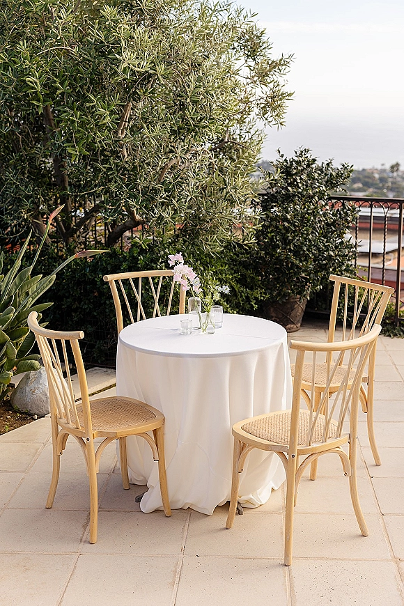 Cocktail table setup with white tablecloth, wooden bistro chairs, bud vases and votive candles on a patio terrace with ocean view