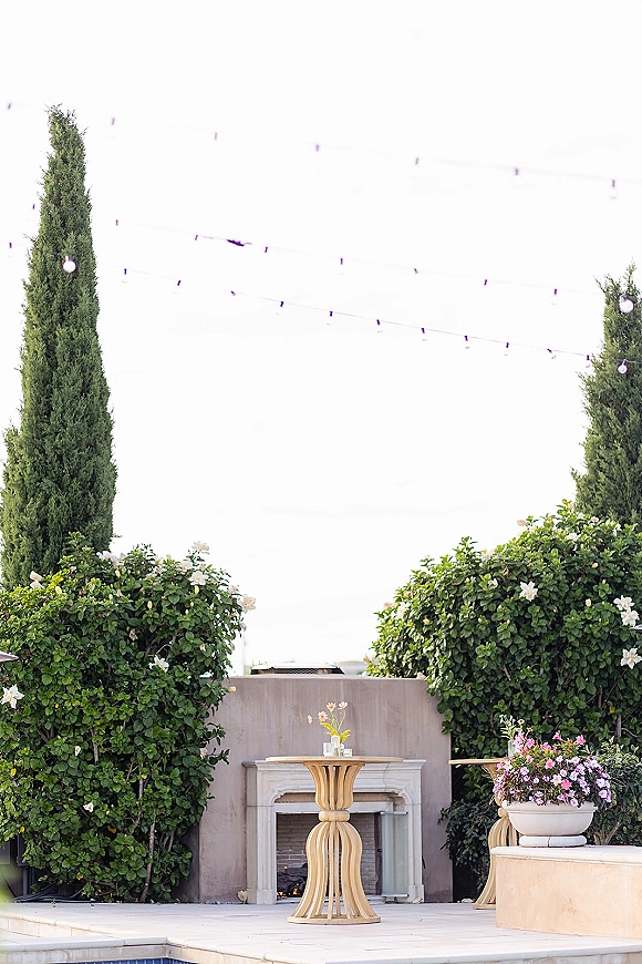 Cocktail hour setup with an outdoor cocktail table, pink and purple floral centerpiece, string lights, and fireplace on a hedge-lined patio