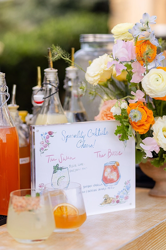 Wedding cocktail bar with signature cocktail sign beside glass drink dispensers, bottles, rocks glasses, and citrus garnish in outdoor greenery